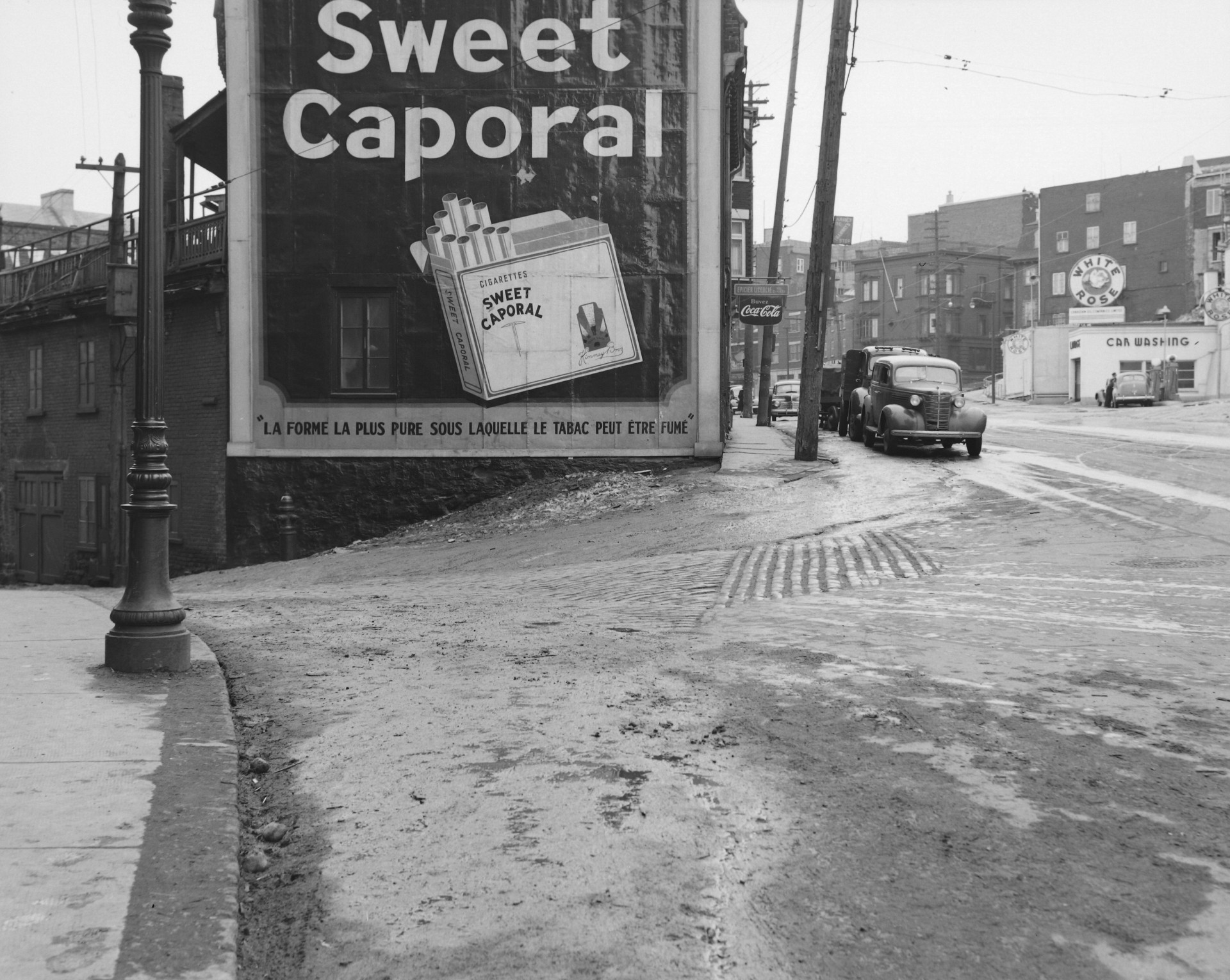 Il s'agit d'un négatif représentant une affiche publicitaire pour les cigarettes Sweet Caporal à l'intersection de la rue St-Olivier et de la côte d'Abraham dans le quartier St-Jean-Baptiste.