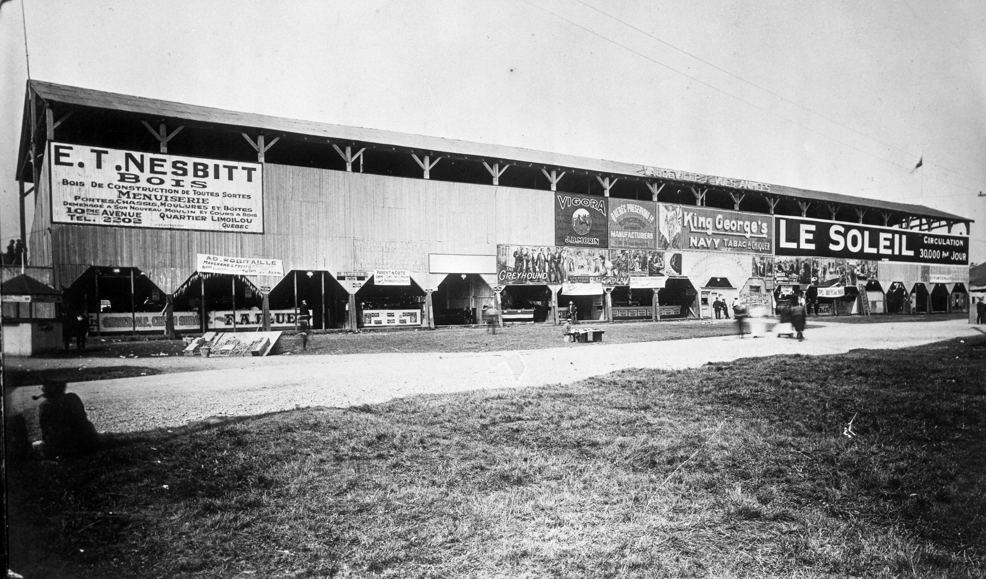 Il s'agit d'une photographie représentant la façade arrière de l'estrade du champs de course au parc de l'Exposition. On remarque des affiches publicitaires, dont celles de E.T. Nesbitt bois, Le Soleil, Vigora J.B. Morin et King George's tabac à chiquer.
